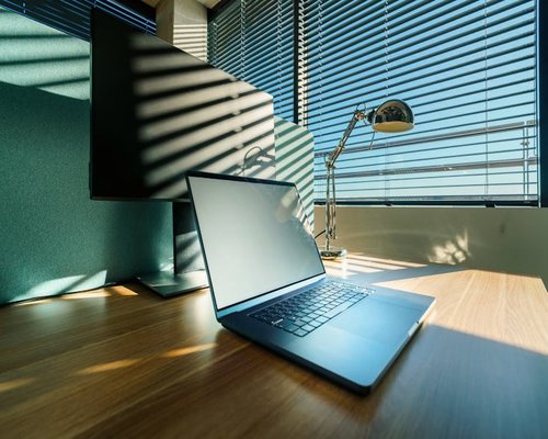 A laptop setup on a wooden desk aligned perpendicular to a large window with soft blinds filtering bright sunlight